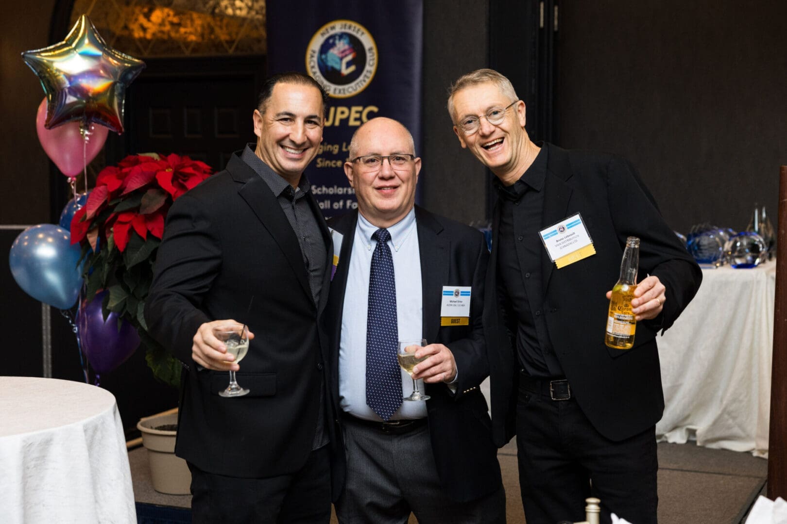 Three smiling men holding drinks at event.